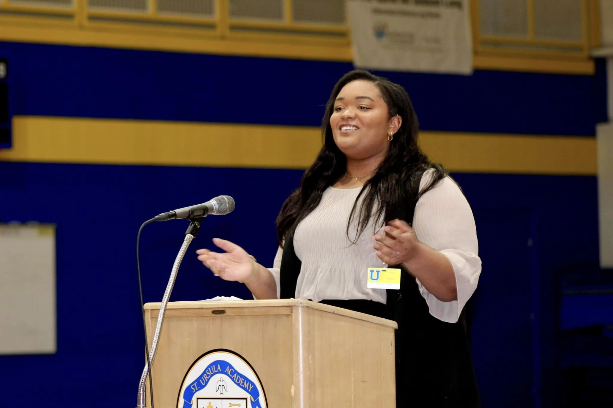 Alayna speaking at a podium marked St. Ursula Academy during a school assembly